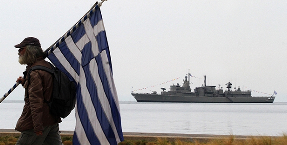 A spectator holds a Greek flag as a Greek Navy vessel is moored at Thermaikos Gulf during the annual military parade in the northern Greek port city of Thessaloniki. (AP Photo/Grigoris Siamidis)