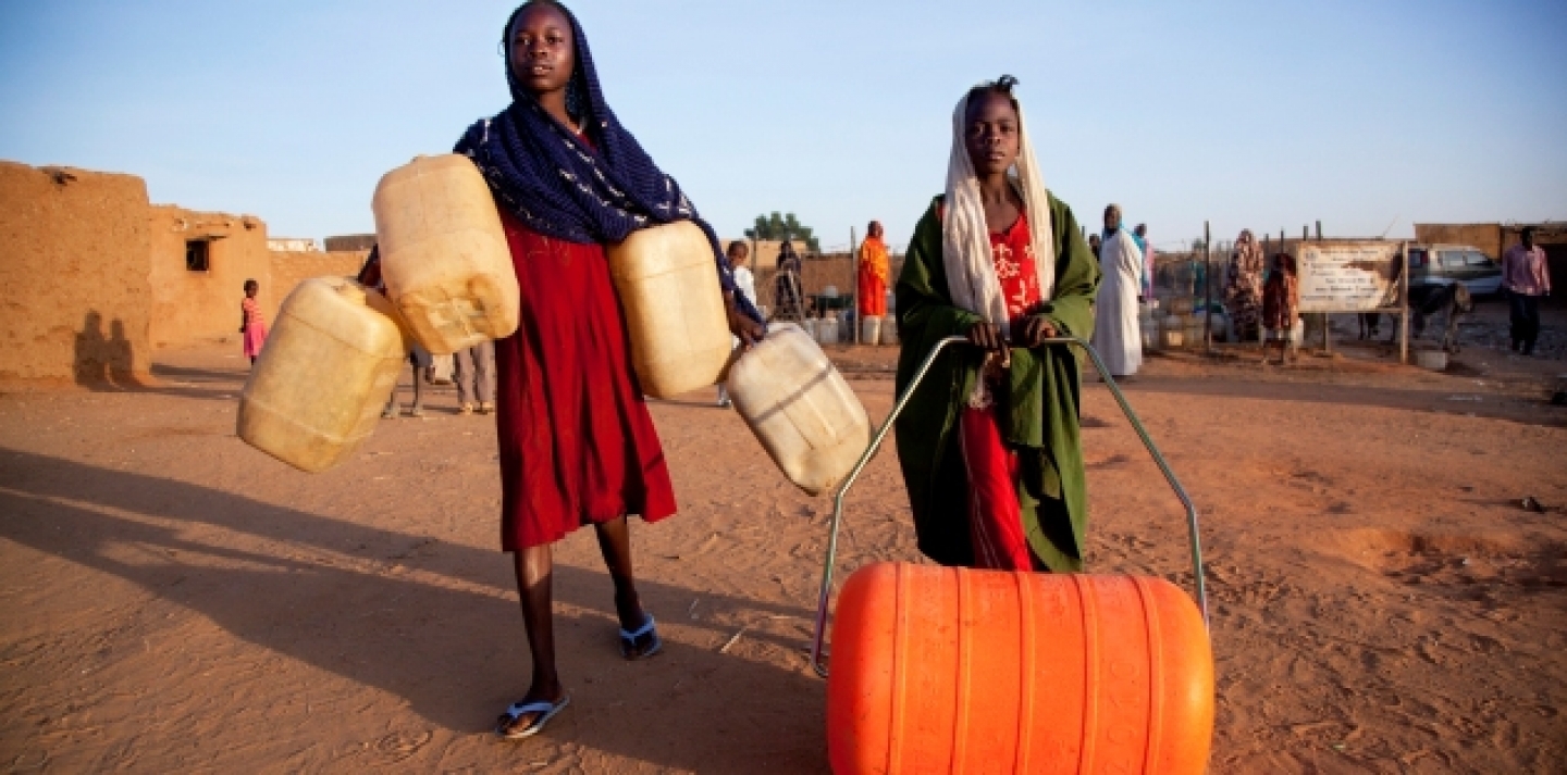 In the Abu Shouk IDP Camp, a child pushes an orange Water Roller, which has the same capacity as the four jerry cans carried by her companion (UN Photo/Albert Gonzalez Farran).
